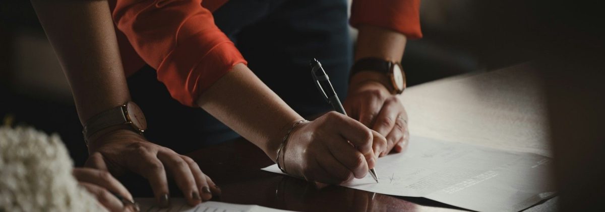 A woman in an orange blazer writes down payment strategies on a piece of paper in a dark office with two others.